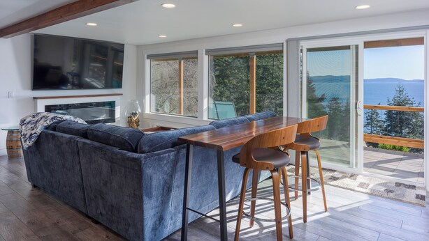 A modern living room with a large sectional sofa, a wooden dining table with bar stools, a flat-screen TV mounted on the wall, and large sliding glass doors leading to a balcony with a view of the sea.