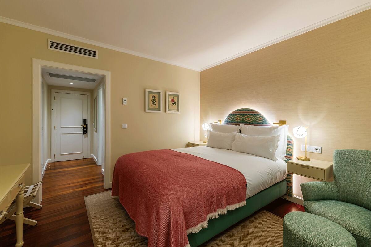 A hotel room with a bed, a nightstand, a green armchair, and a door leading to another room.