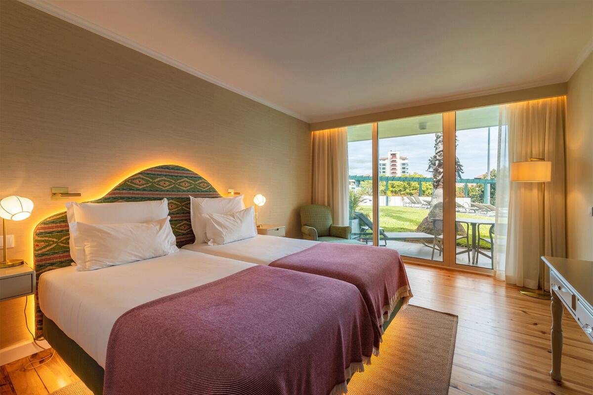 A hotel room with two beds, a large window with a view of the outdoors, and a balcony with outdoor seating.