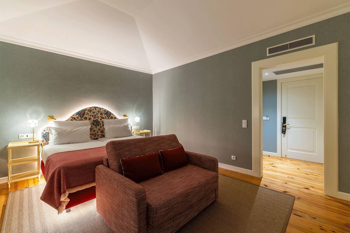 A bedroom with a bed, a red armchair, a nightstand, and a door.