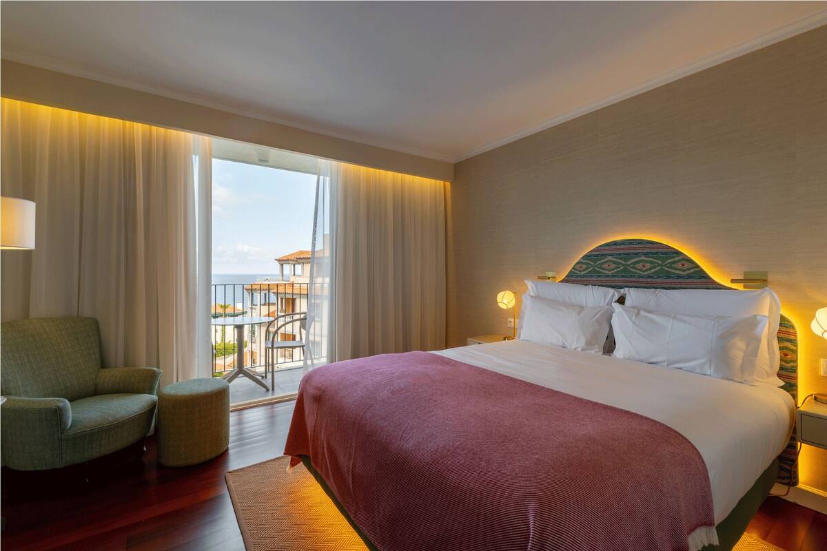 A hotel room with a large bed, a green armchair, a small ottoman, and a balcony with a view of the sea.