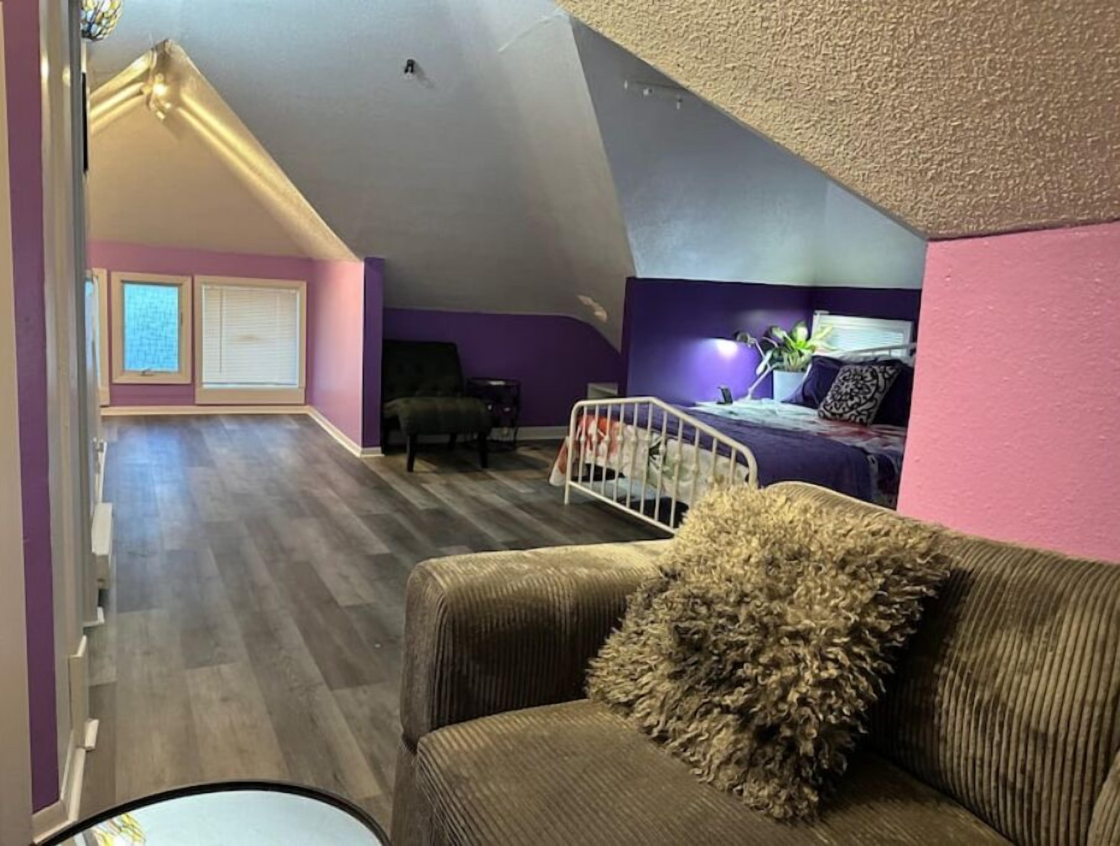 A spacious loft bedroom with a sloped ceiling, a large window, a bed, and a seating area with a couch and chair.