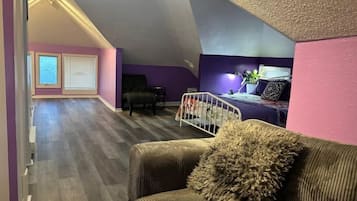 A spacious loft bedroom with a sloped ceiling, a large window, a bed, and a seating area with a couch and chair.
