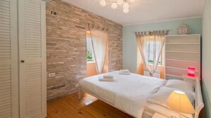 A bedroom with a wooden floor, a bed with white linens, a nightstand with a lamp, and a wall with a brick pattern.