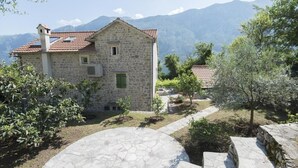A stone building with a red-tiled roof, surrounded by greenery and mountains in the background.
