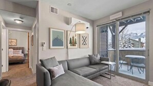 A modern living room with a grey sofa, a small table, and a balcony with a view of snow-covered mountains.