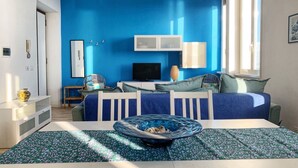 Une pièce avec un mur d’accent bleu, un lit avec une literie bleue, une télévision, une armoire blanche et une table à manger recouverte d’une nappe à motifs.