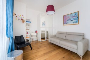 A living room with a white sofa, a black chair, a small table, a blue curtain, and a painting on the wall.