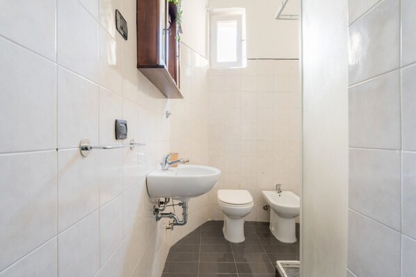 A small bathroom with white tiles, a sink, two toilets, and a bidet.