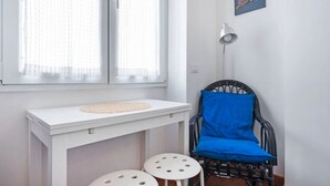Et værelse med et hvidt bord, to barstole, en sort fletstol med blåt sædehynde, et vindue med lette gardiner og en vægmonteret lampe.