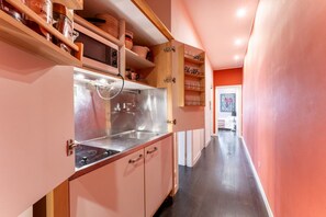 A narrow hallway with a kitchen area featuring pink cabinets, stainless steel appliances, and wooden shelves.