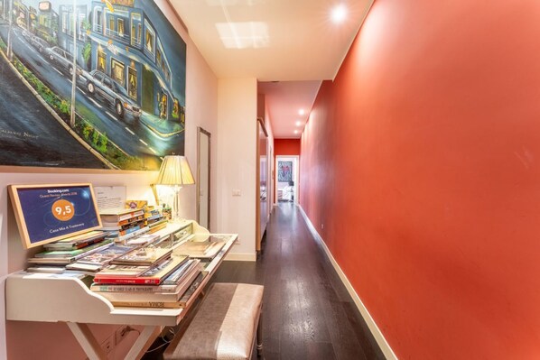 A hallway with a desk displaying books and a framed certificate, a painting of a street scene, and a red wall.