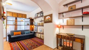 A living room with a sofa, a patterned rug, a dining table with chairs, and shelves with decorative items.