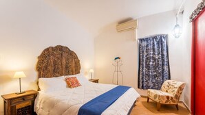 A bedroom with a large bed, a chair, a nightstand, a wall-mounted air conditioner, and a window with a patterned curtain.