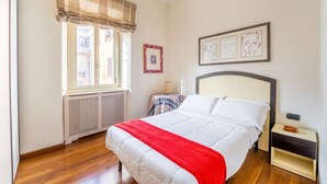 Un dormitorio con una cama, un radiador, una ventana con un visillo a rayas y obras de arte enmarcadas en la pared.
