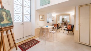 Une pièce comprenant une table à manger, des chaises, un tableau sur chevalet et une grande fenêtre ornée d’un motif géométrique.