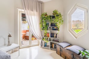 A room with a white desk, a bookshelf with plants, a window with a view, and a wooden chest.