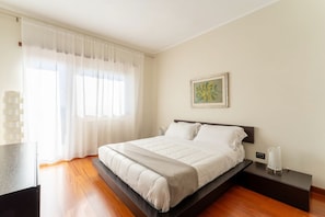 A modern bedroom with a bed, bedside tables, a nightstand, and a window with sheer curtains.