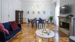A room with a blue sofa, a round table with candles, a dining table with chairs, a fireplace, and a mounted TV. - GLAM 50 - Exclusive Penthouse in the Heart of Rome (Roma)