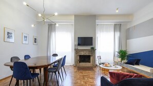 A modern living room with a fireplace, a dining area with a round table and chairs, a TV mounted on the wall, and a sofa with cushions. - GLAM 50 - Exclusive Penthouse in the Heart of Rome (Roma)