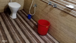 Um banheiro com vaso sanitário, chuveiro e um balde vermelho no chão.