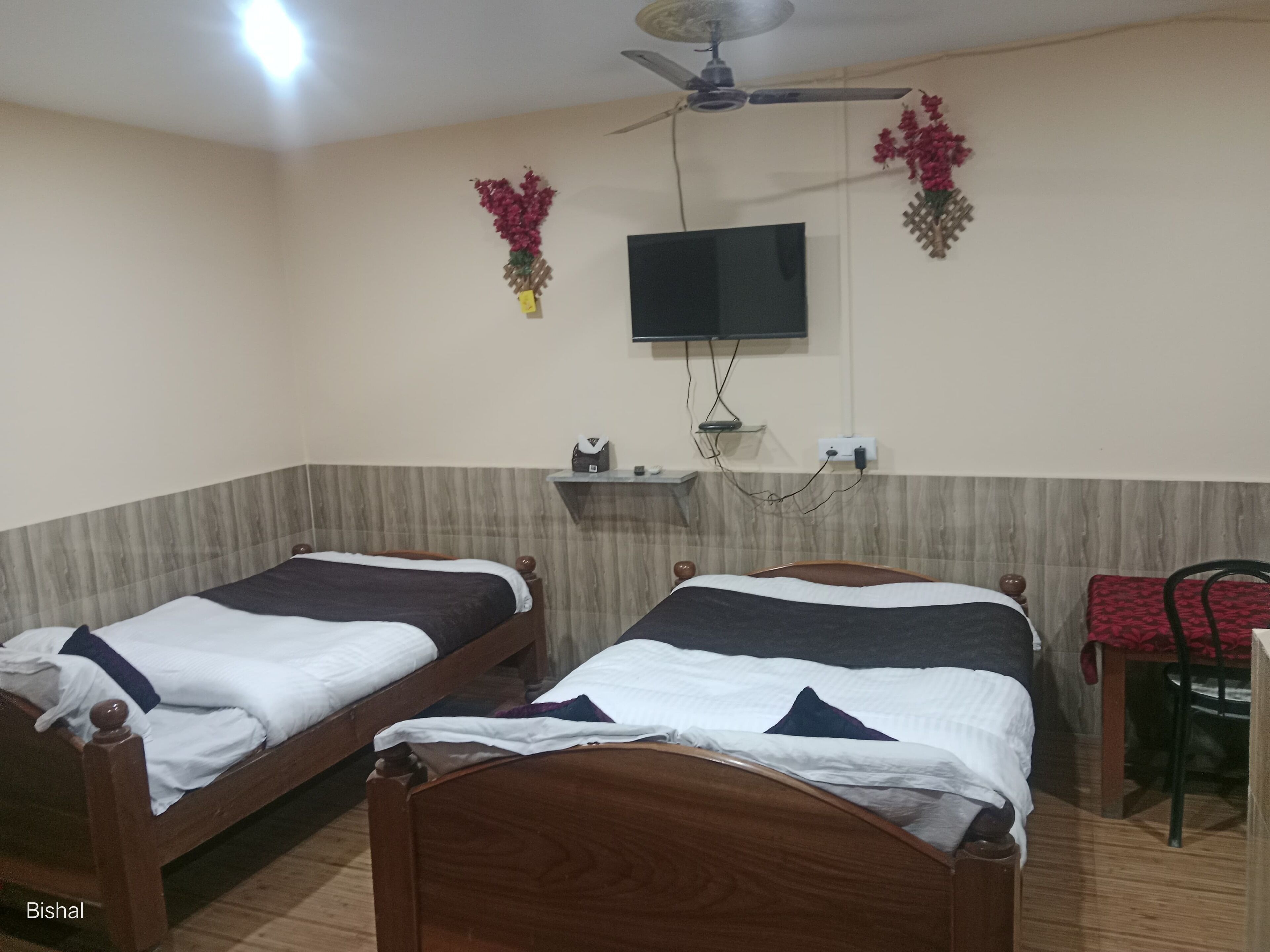 A hotel room with two wooden beds, a TV mounted on the wall, a ceiling fan, and a small shelf with a vase of flowers.
