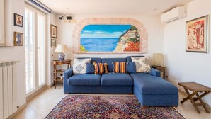 Una sala de estar con un sofá, un tapete estampado, un cuadro de una escena costera y una mesita.