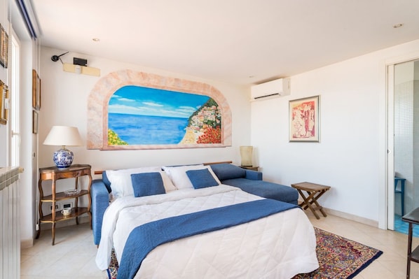 A bedroom with a bed, a painting of a beach, a small table, and a chair.