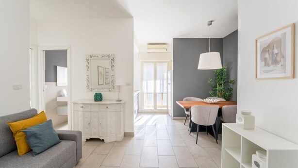 Una sala de estar moderna con un sofá gris, un armario blanco y un comedor con mesa y sillas.