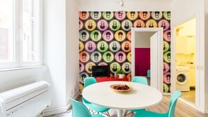 Ett kompakt kök med ett färgstarkt väggmönster, en matplats med turkosa stolar och en tvättmaskin som syns genom en öppen dörr.
