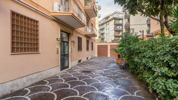Un vicolo stretto con pavimentazione decorata, un edificio con una finestra sbarrata e un siepe verde.