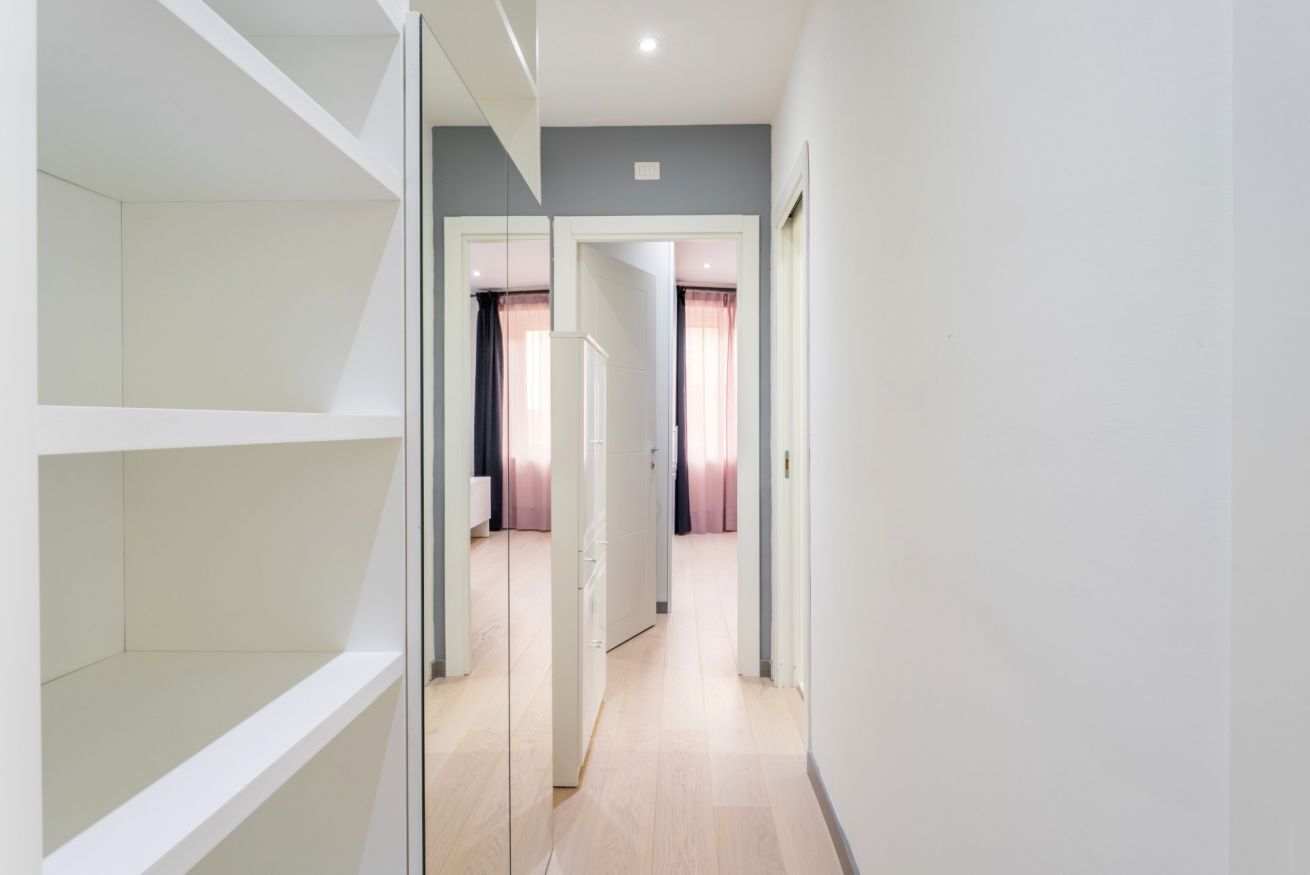 Un corridoio con un armadio bianco, una porta che dà su una stanza con un letto e tende, e un pavimento chiaro.