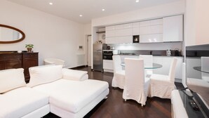 A modern living room with a sofa, chairs, a glass coffee table, a kitchen area with cabinets, a refrigerator, and a clock. - The Vatican Rest, Elegant Apt Nerby the Basilica (Roma)