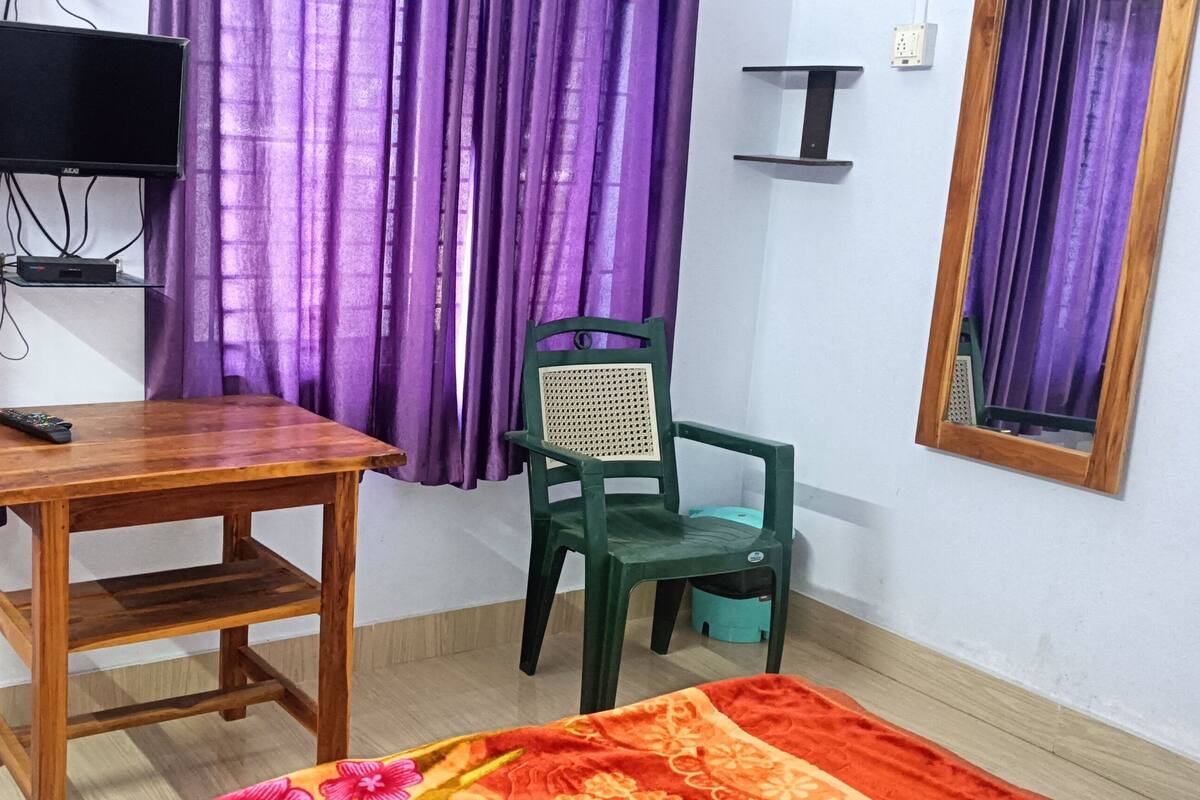Une pièce avec une table en bois, une chaise verte, un téléviseur fixé au mur, des rideaux violets et une petite étagère.