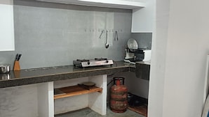 A compact kitchen with a gas stove, hanging utensils, and a gas cylinder. - Sneha Homestay by Roh (Bokakhat)
