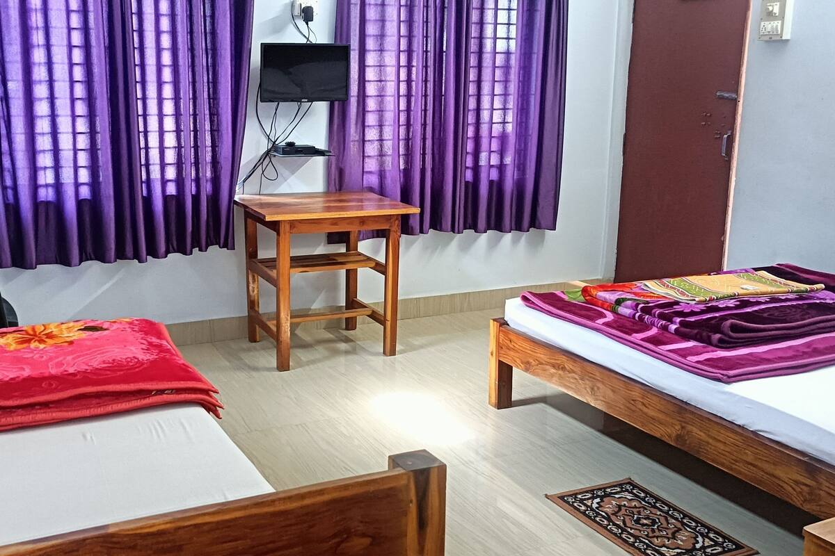 Une petite pièce bien rangée, avec deux lits, une table en bois, une chaise et des rideaux violets.