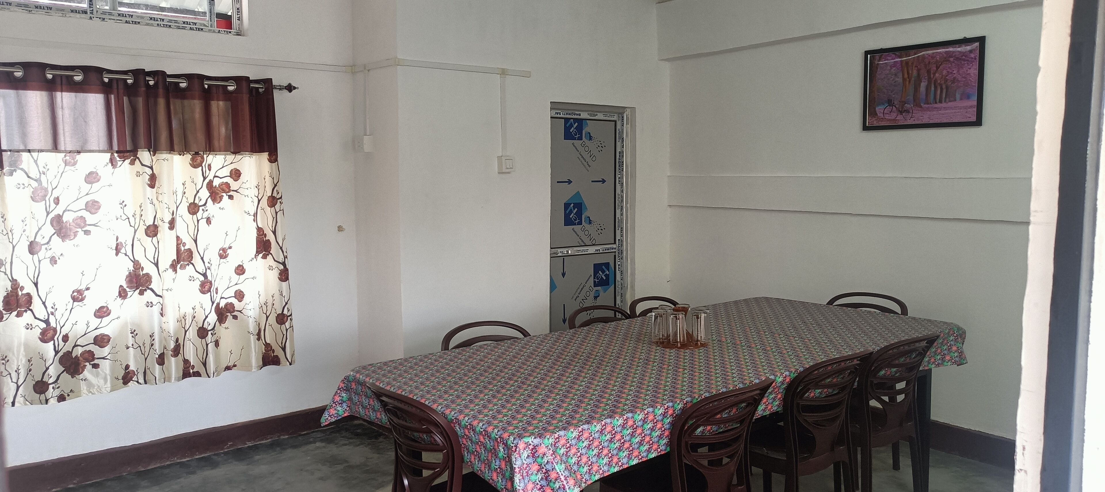 A dining area with a table set for four, surrounded by chairs, a window with floral curtains, and a wall-mounted picture.