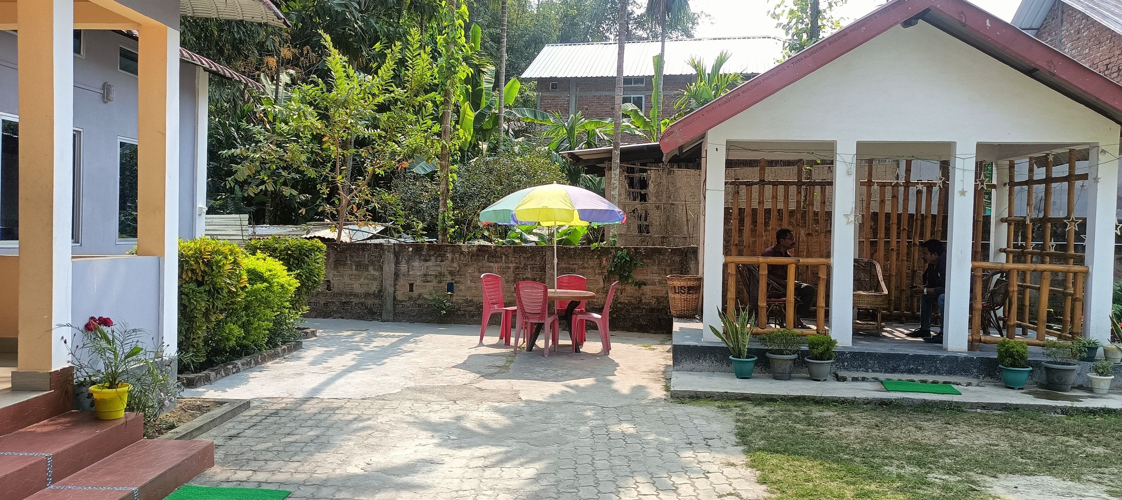 A courtyard with a table and chairs under a colorful umbrella, surrounded by potted plants and a small building with bamboo fencing.