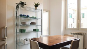 A dining area with a wooden table and chairs, a metal shelving unit with dishes and plants, and a view of buildings through a window. - Casa Vic Garbatella in Rome (Rome)