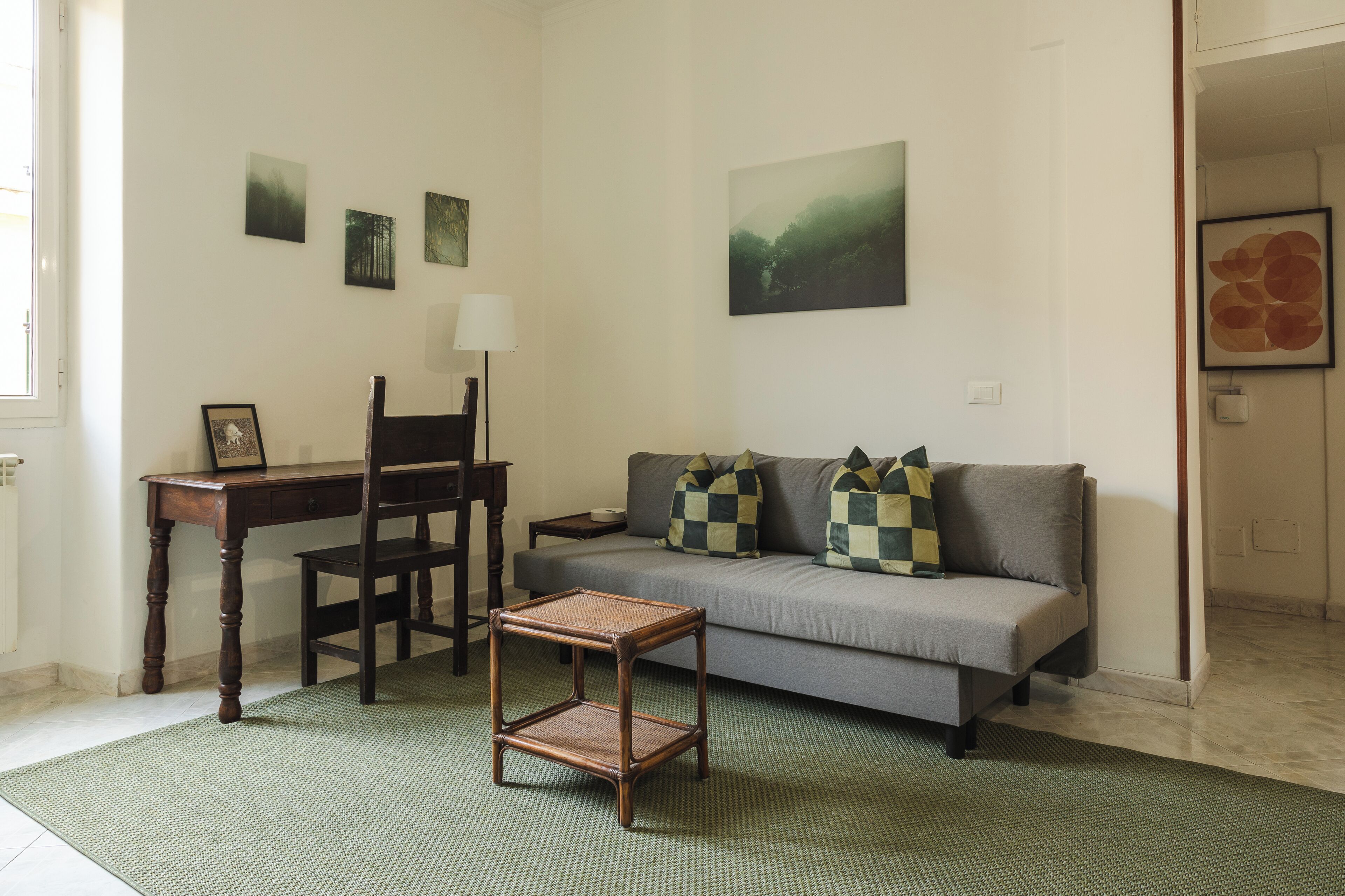 A living room with a sofa, a small table, a chair, and a lamp. There are three framed pictures on the wall.
