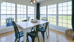 Uma área de refeições com uma mesa branca e cadeiras verdes, grandes janelas com vista para o mar e um lustre.