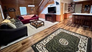 Ein geräumiges Wohnzimmer mit einer großen Couch, einem Couchtisch und einem gemusterten Teppich. Eine Treppe führt zu einer oberen Ebene mit einem Fernseher und Regalen.