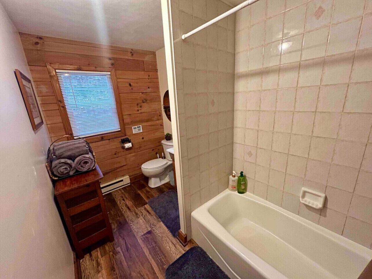 A bathroom with a wooden floor, a white bathtub, a toilet, and a window with blinds.