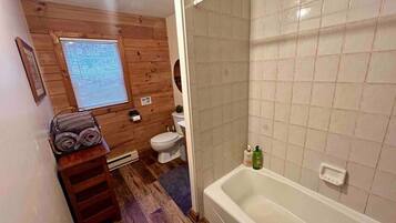 A bathroom with a wooden floor, a white bathtub, a toilet, and a window with blinds.