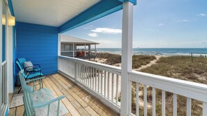 Eine überdachte Veranda mit blauen Wänden und einem Holzboden, die auf einen Strand mit einem Dünenweg und einem Boot in der Ferne blickt.