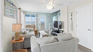 A living room with a ceiling fan, a television, and a beach-themed wall art. - AOK Oasis House by Liquid Life (Fort Morgan)