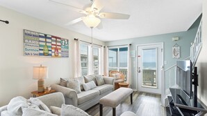 A living room with a ceiling fan, a sofa set, a wicker chair, a TV, and a window with blinds. - AOK Oasis House by Liquid Life (Fort Morgan)