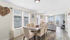A spacious dining area with a wooden table and chairs, large windows with blinds, and a ceiling fan. - AOK Oasis House by Liquid Life (Fort Morgan)