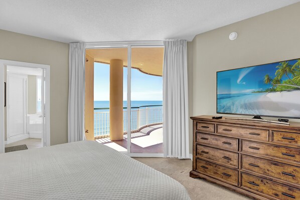 Habitación de hotel con una cama grande, un aparador de madera, un televisor y un balcón con vista al océano.