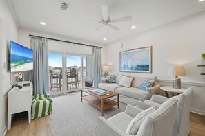 A spacious living room with a large flat-screen TV, a ceiling fan, and a view of the ocean through the sliding glass doors.